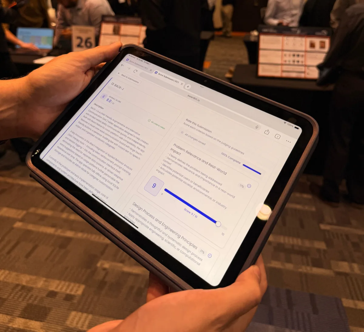 A judge at ECS Innovation Expo 2026 scoring a project on an iPad using AwardKit, with project posters and other judges in the background
